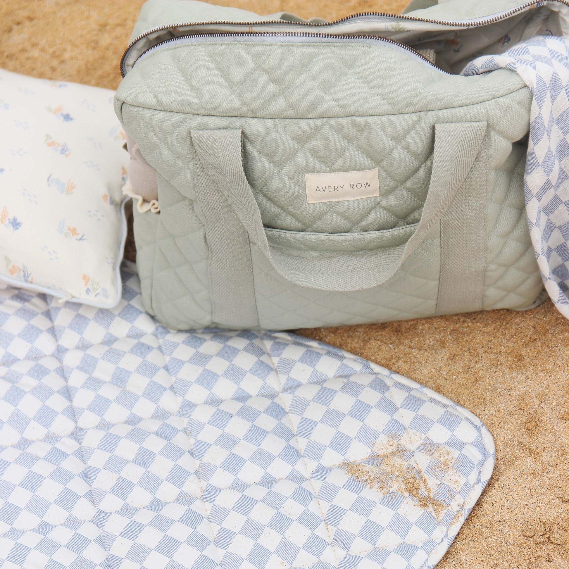 Green quilted bag with a visible brand logo on a sandy surface with a checkered blanket.