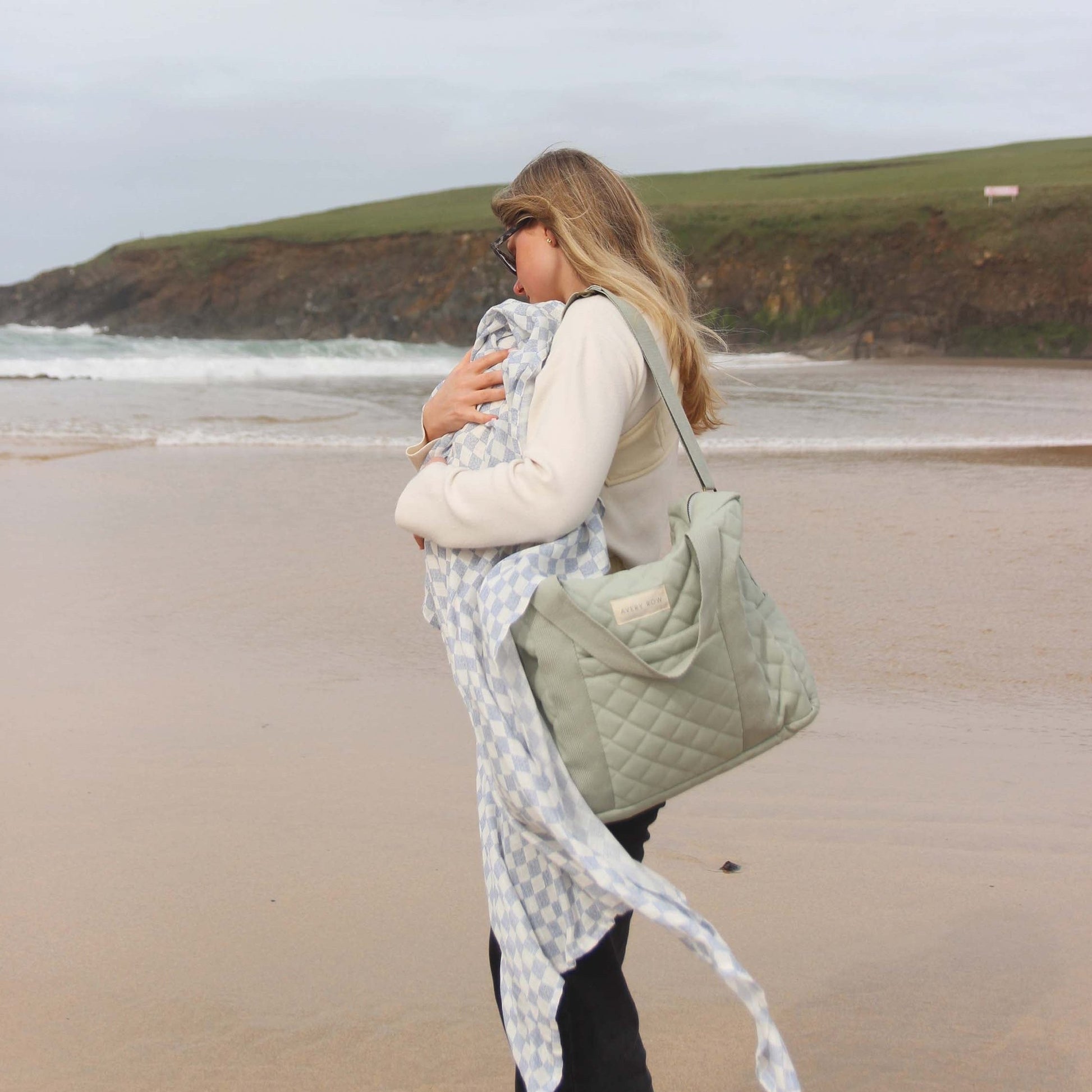 Woman holding a baby and a green bag on a beach with a scenic background