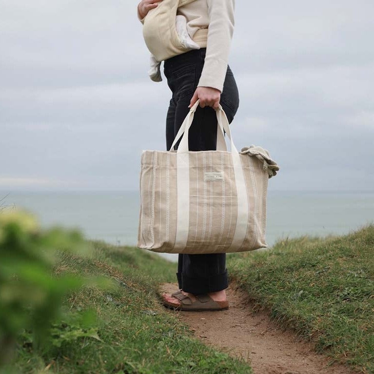 Person holding a large beige tote bag outdoors near a body of water.