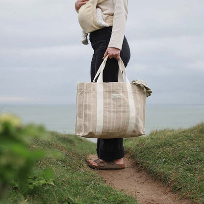 Person holding a large beige tote bag outdoors near a body of water.