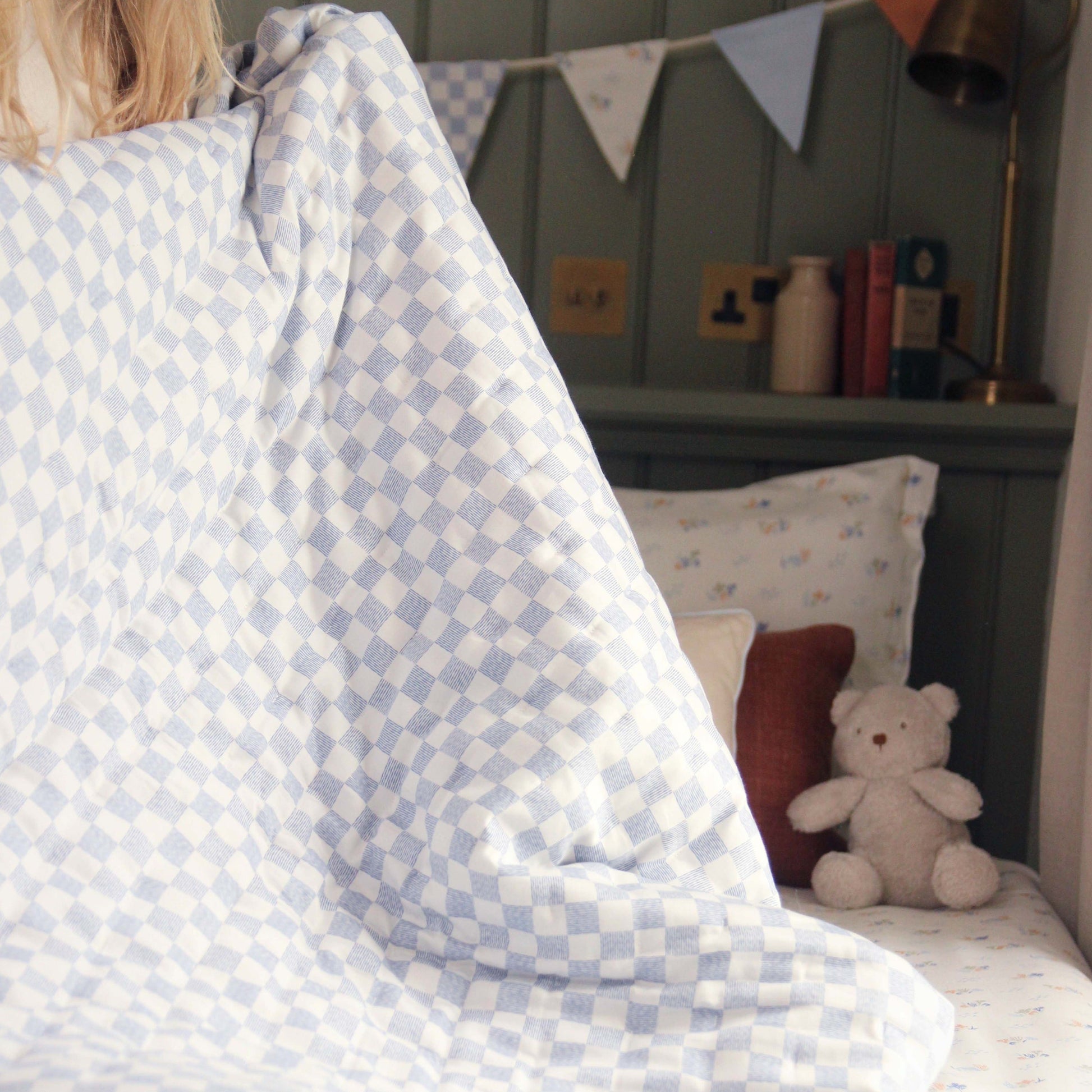Child under a blue and white checkered blanket with a teddy bear and pillows in the background.