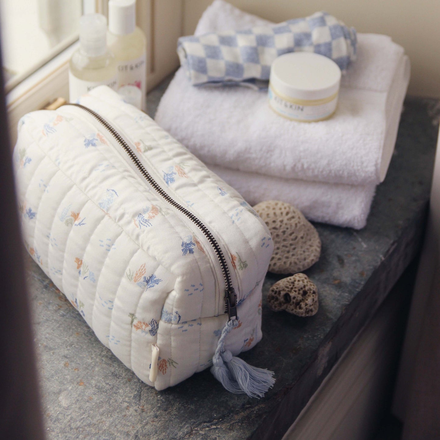 Quilted toiletry bag with floral pattern on a bathroom counter with towels and bottles.