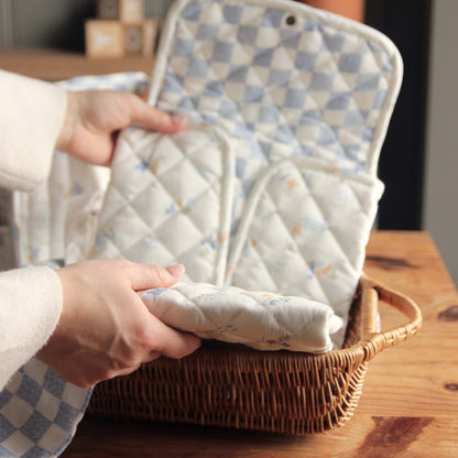 Person holding a quilted baby blanket over a wicker basket on a wooden surface.