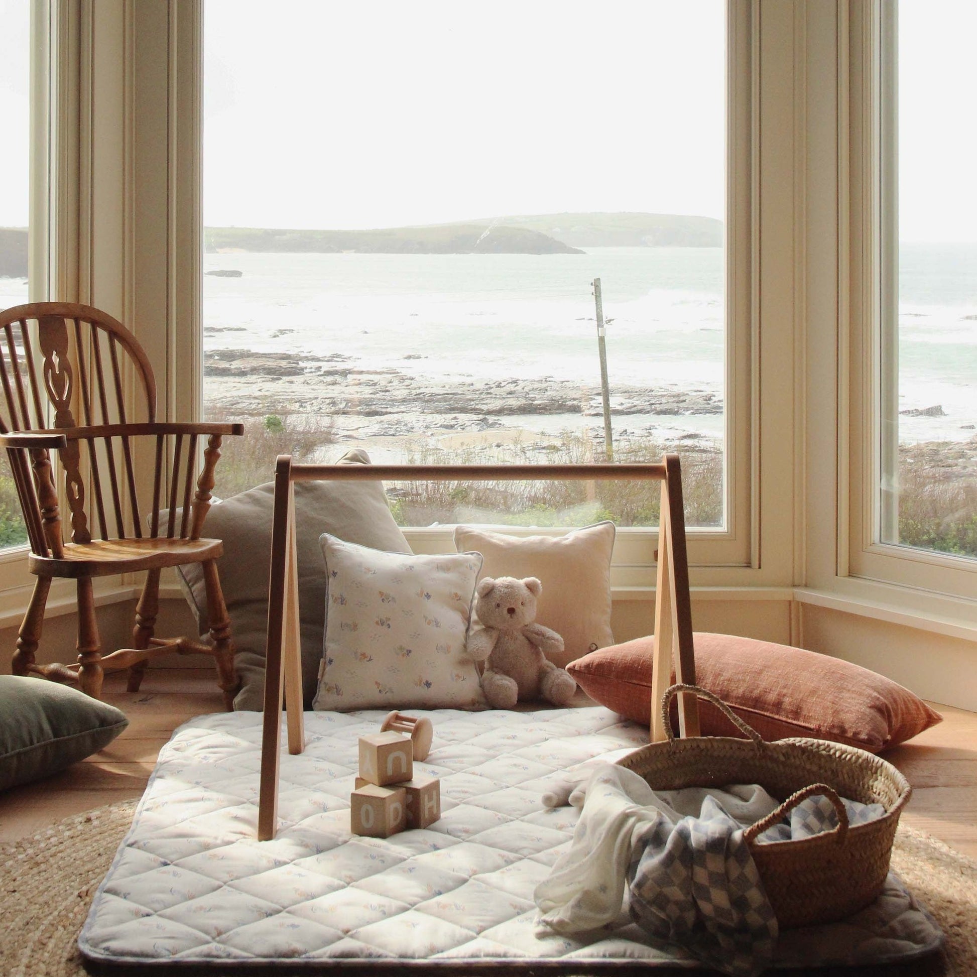 Children's play area with a wooden frame, cushions, a teddy bear, and toys in a room with large windows.