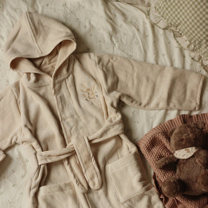 Beige robe with floral embroidery on a textured surface with a brown knitted item and teddy bear.