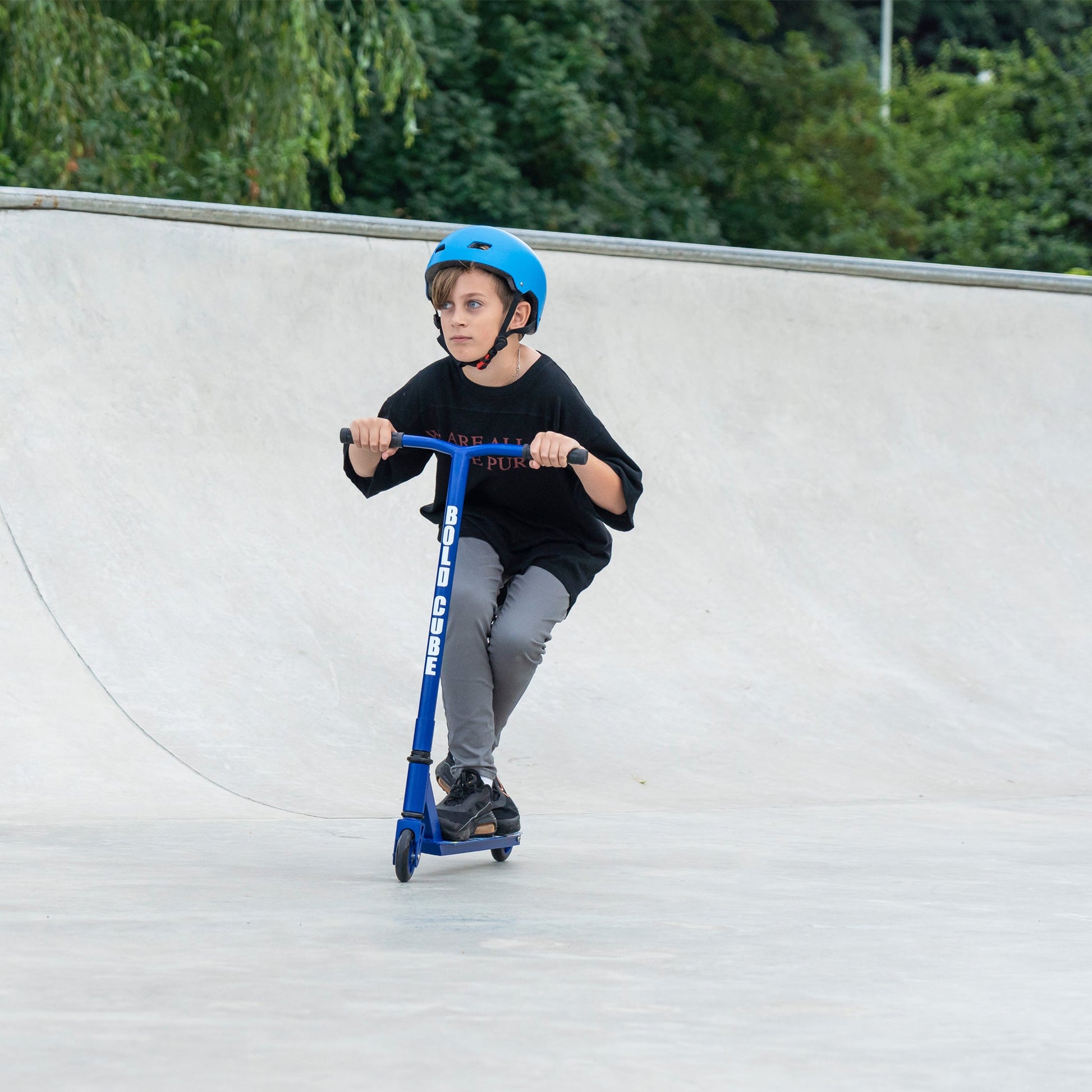 Child riding a blue scooter at a skate park with greenery in the background