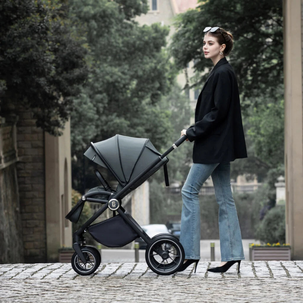 Woman pushing a black stroller on a paved path with greenery in the background