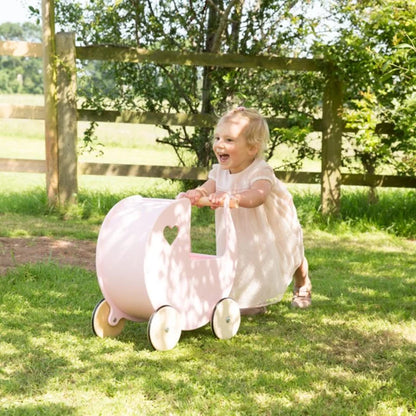 Child in a white dress playing with a pink toy cart in a grassy outdoor setting.