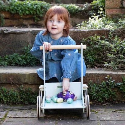 Child sitting in a toy stroller with a purple teddy bear and colorful eggs outdoors.