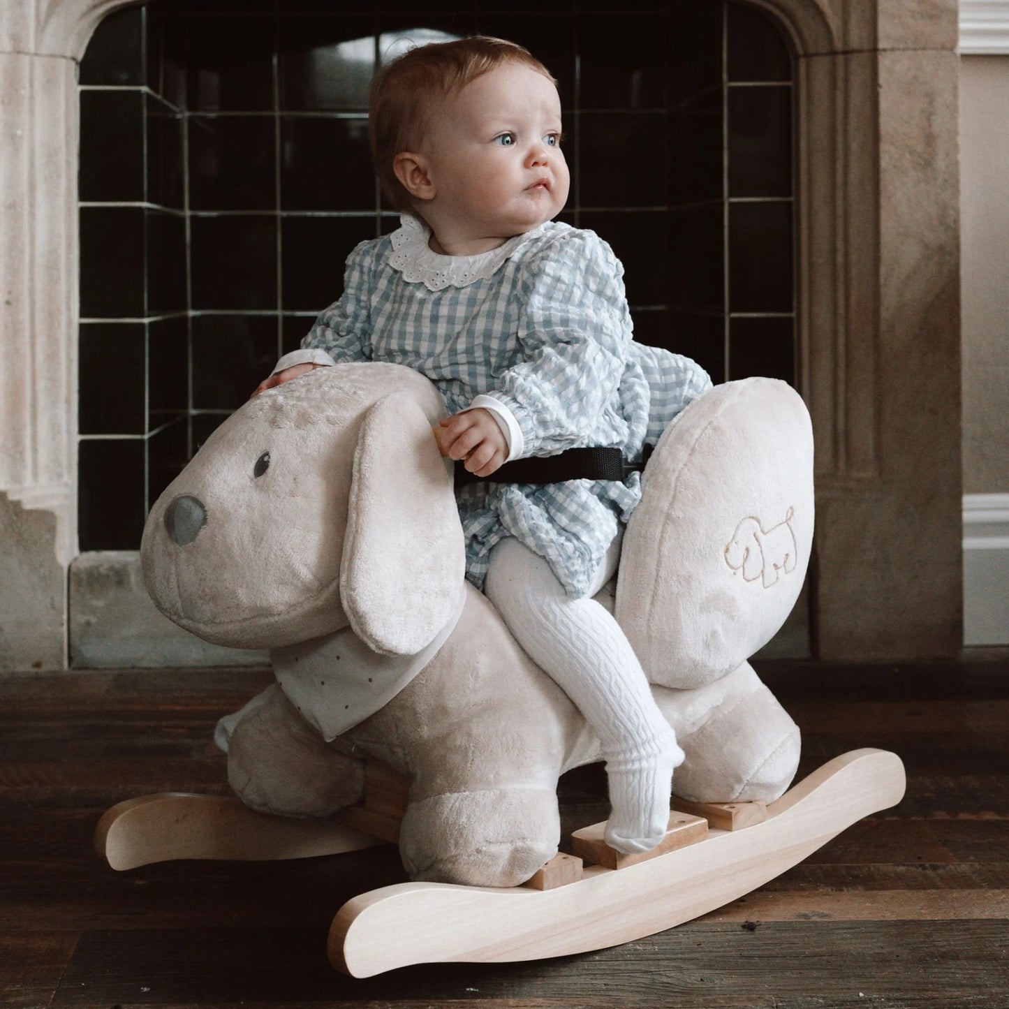 Baby sitting on a rocking horse in a room with tiled walls.
