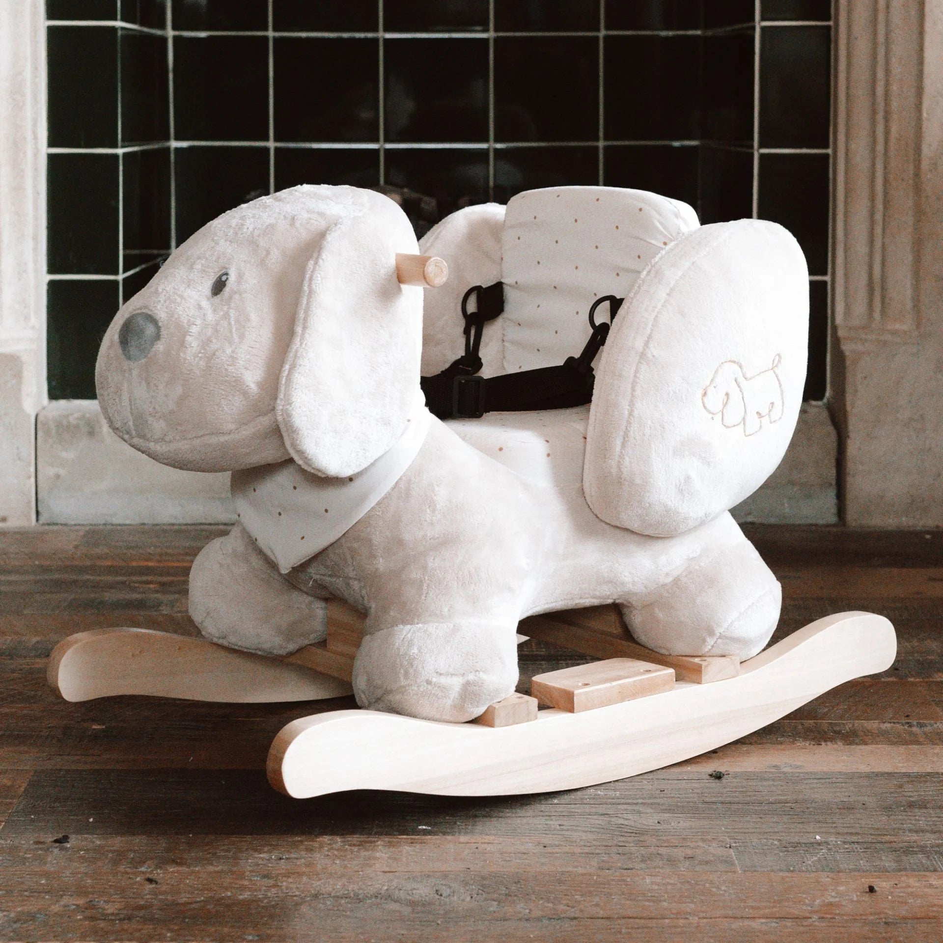 Plush dog-shaped rocking toy on a wooden floor with a tiled wall in the background