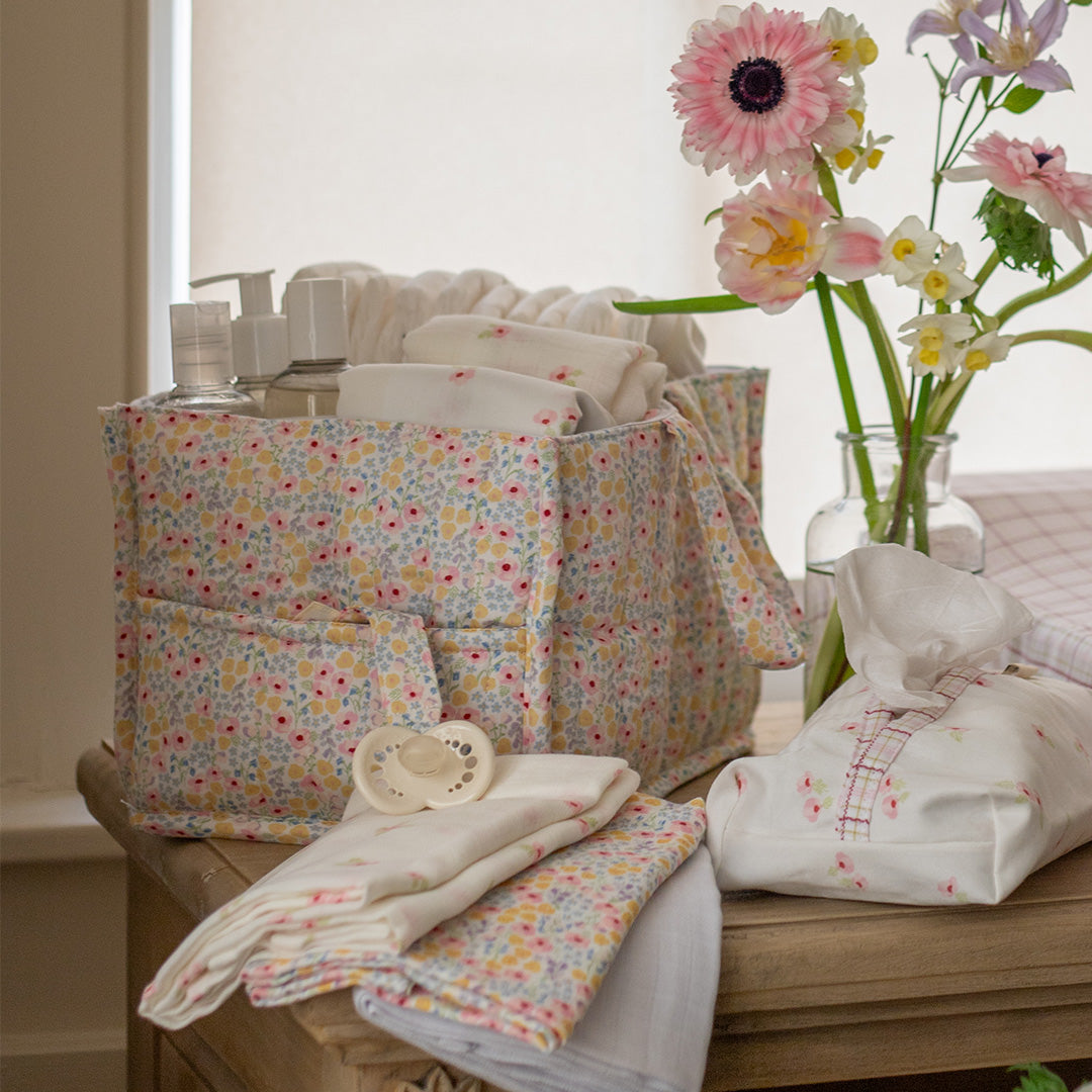 Floral-patterned diaper bag with baby items on a wooden surface, next to a vase of flowers.