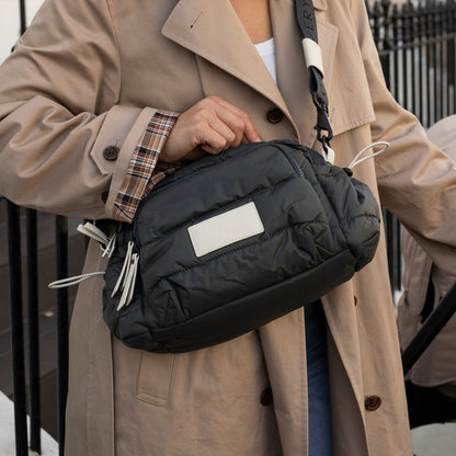 Person holding a black bag with a visible brand label, wearing a beige coat.