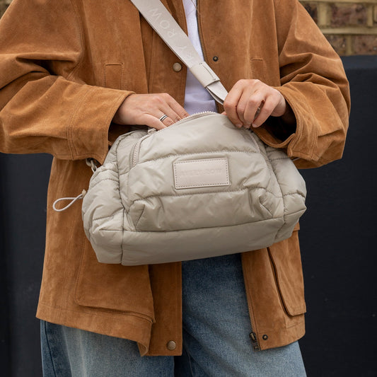 Person wearing a brown jacket holding a beige quilted bag against a dark background