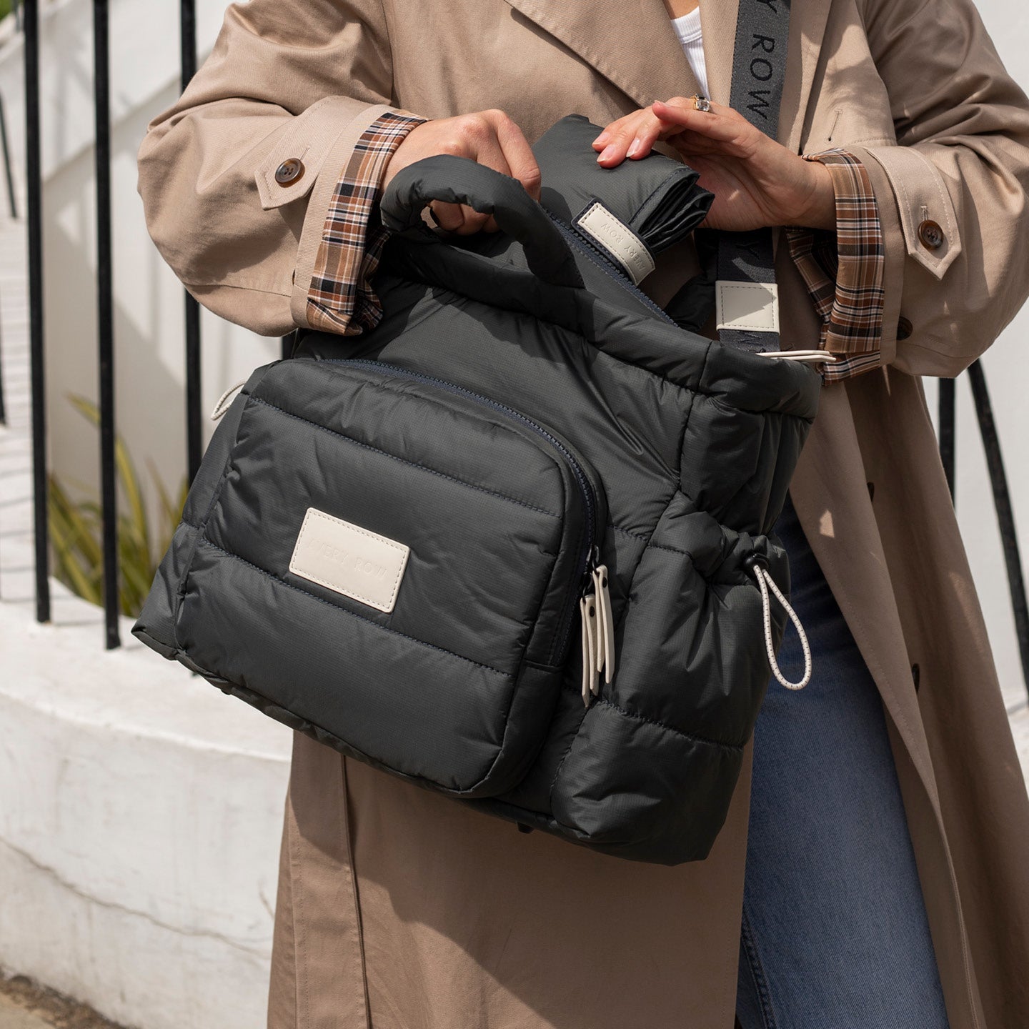 Person holding a black puffer bag with a brand label, wearing a beige coat.