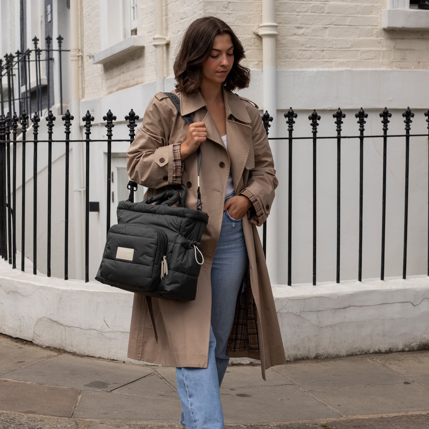 Woman in a beige coat holding a black backpack on a city street.