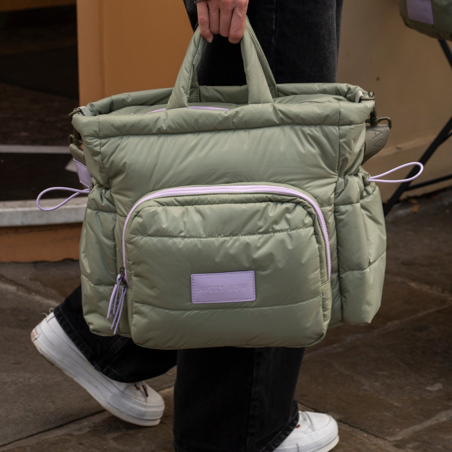 Green puffer bag with a purple label held by a person wearing white sneakers.