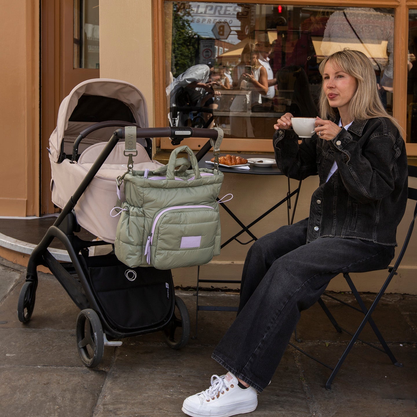 Woman sitting with a coffee cup next to a stroller with a green bag in an outdoor cafe setting.