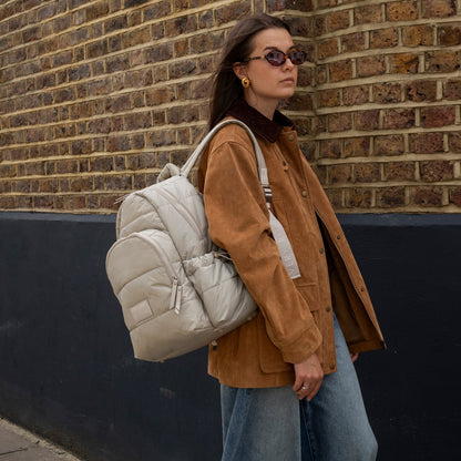 Woman wearing a beige quilted backpack against a brick wall.