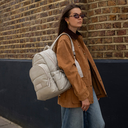Woman wearing a beige quilted backpack against a brick wall.