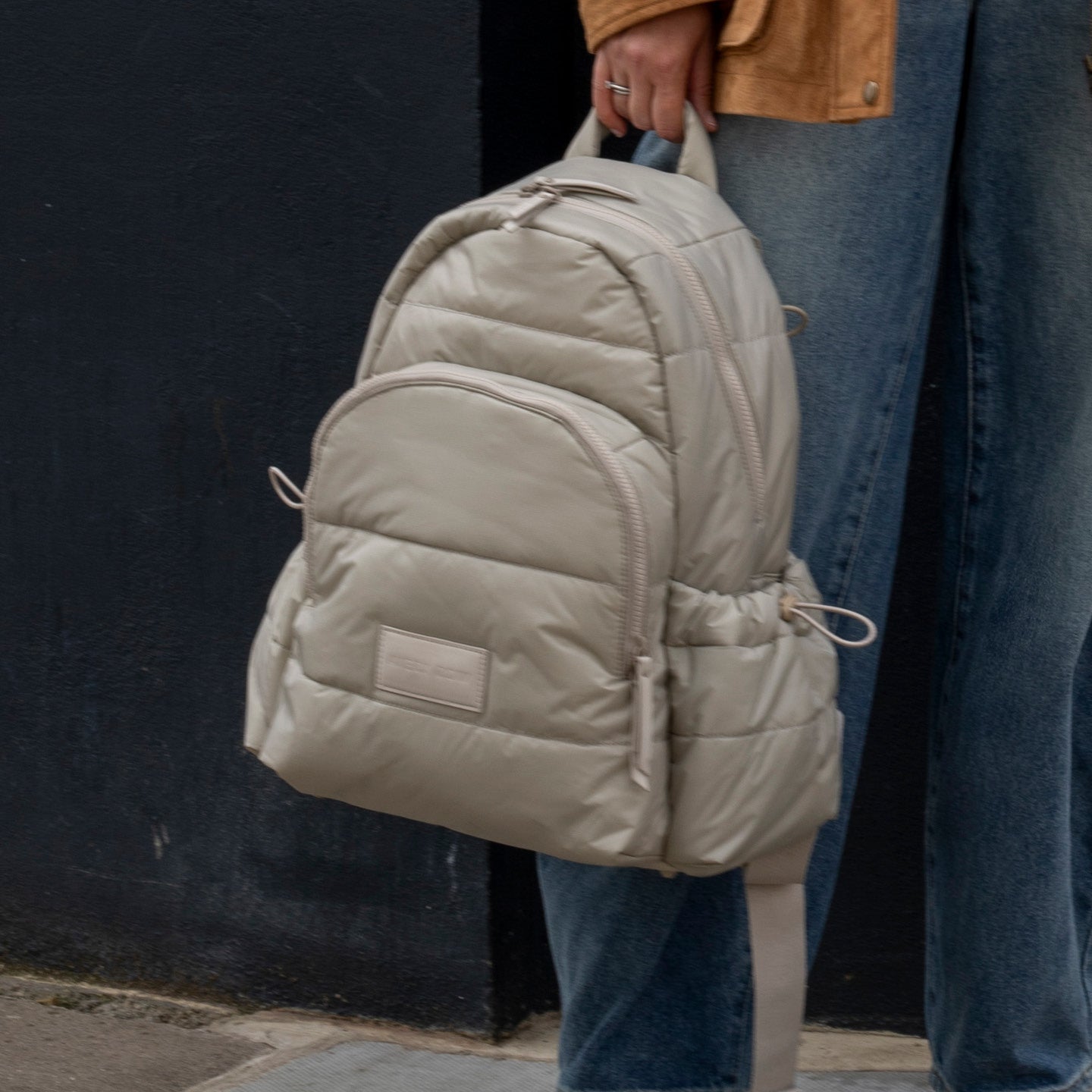 Beige puffer backpack held by a person wearing blue jeans and a brown jacket.