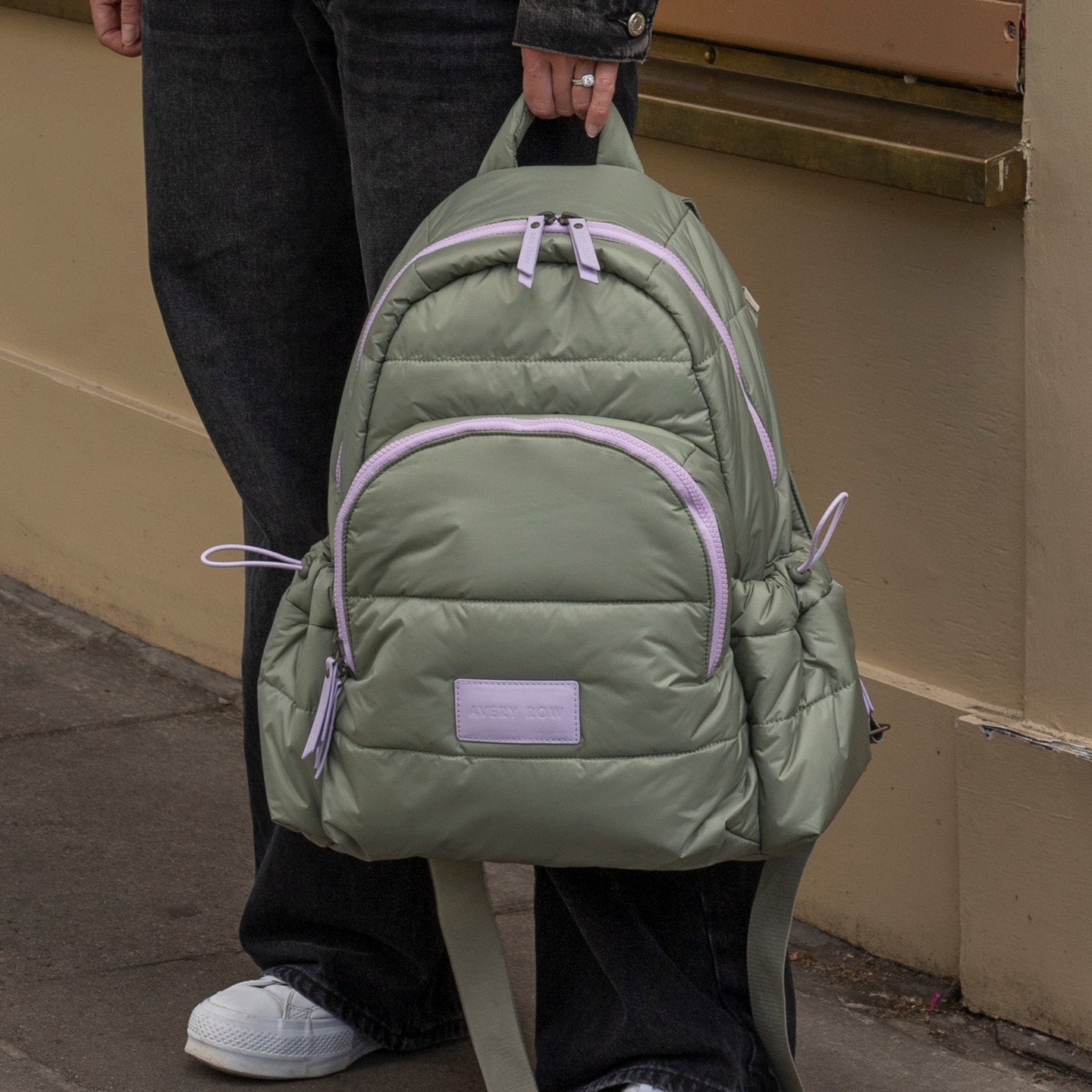Person holding a green backpack with pink zippers against a beige wall.