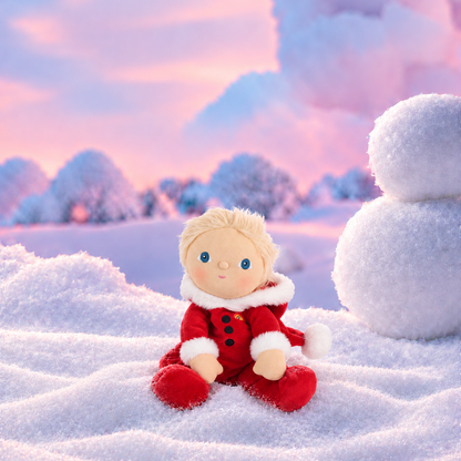 A small blonde doll wearing a red outfit with white trim sits in a snowy landscape. The background shows a pink and blue sky, snow-covered trees, and a large snowball on the right.