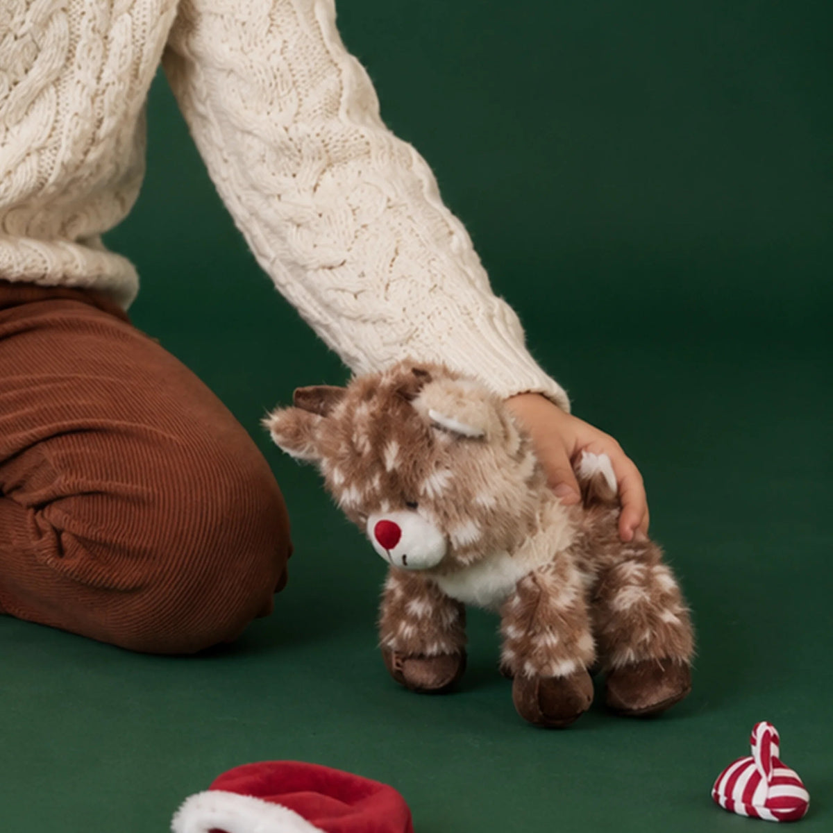 A small reindeer plush toy with antlers and a red nose on a green background with a small childs knee and arm in picture.
