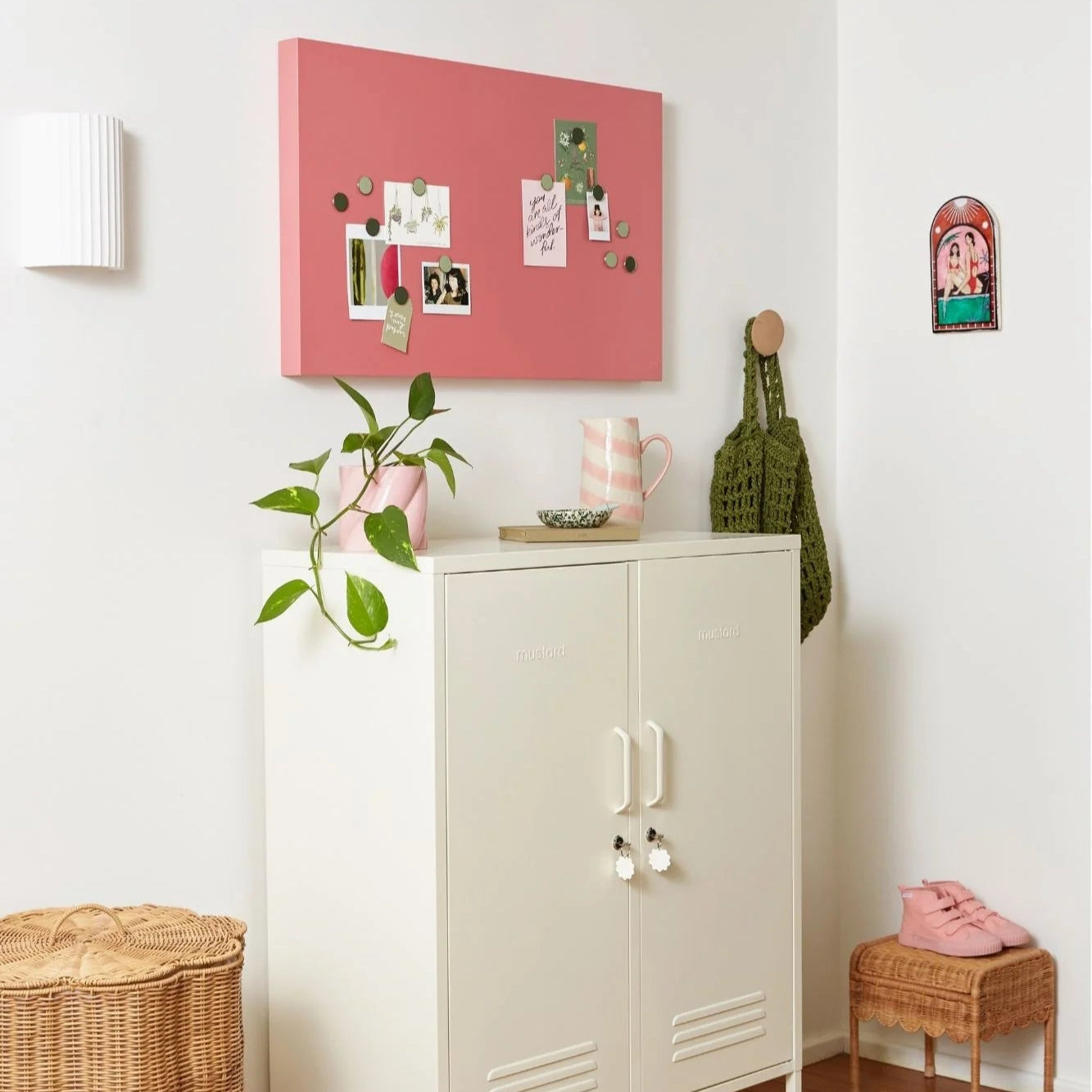 White cabinet with pink bulletin board above, plant to the left, and small table with pink bag to the right.