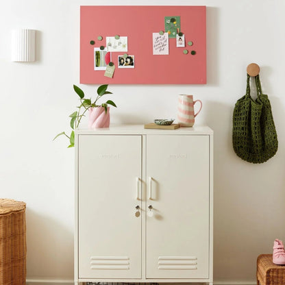 White cabinet with pink bulletin board above, plant to the left, and green bag on the right.