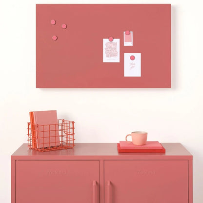 Pink cork bulletin board with pins on a white wall above a pink locker.