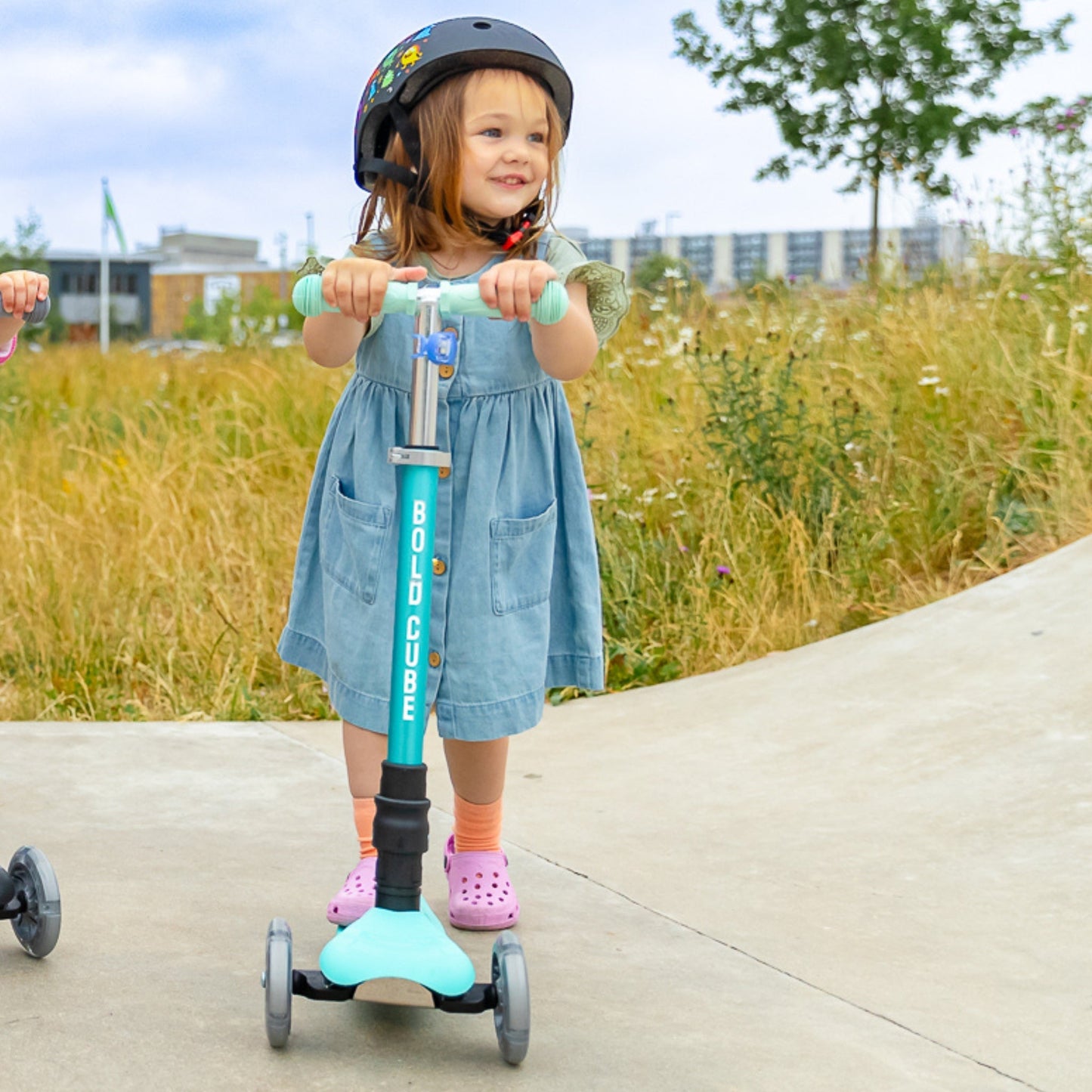 Child riding a scooter with a helmet on a sidewalk with grass and buildings in the background