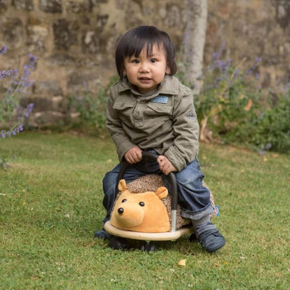 Child riding a hedgehog-shaped toy on grass with a stone wall and flowers in the background