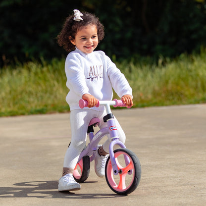 Child riding a balance bike outdoors on a paved path with grass and trees in the background.