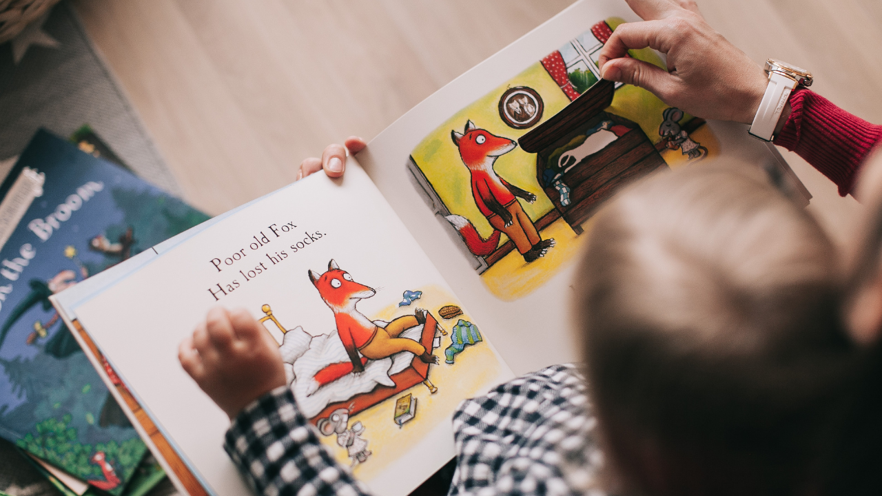 Child reading a book with a fox illustration, surrounded by other books on a wooden floor.