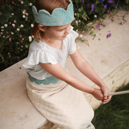 Child wearing a light blue crown and white outfit with ruffled sleeves, sitting outdoors.