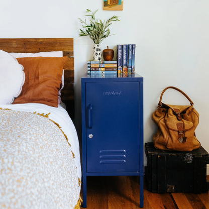 Blue metal locker against a white wall with a bed, plant, and hat in the background.