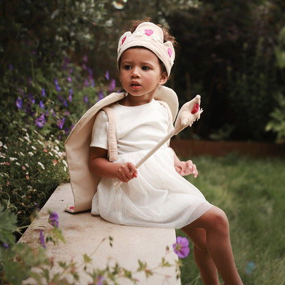 Child in a white dress with bunny ears and a wand, sitting outdoors with flowers in the background