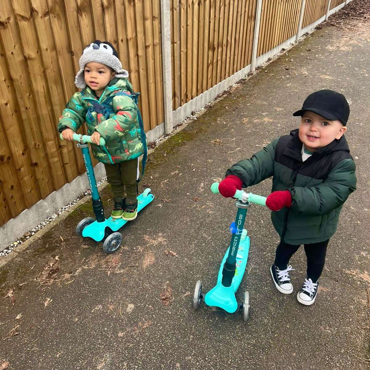 Two children with turquoise scooters on a paved path next to a wooden fence.