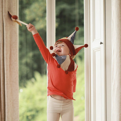 Child wearing a colorful elf hat and red shirt standing by a window with greenery outside.