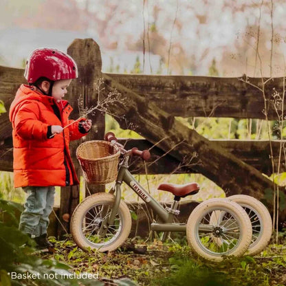 Child in red jacket and helmet standing next to a green balance bike with basket not included, outdoors.