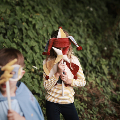 Person wearing a jester hat and mask holding a staff in front of a green bush