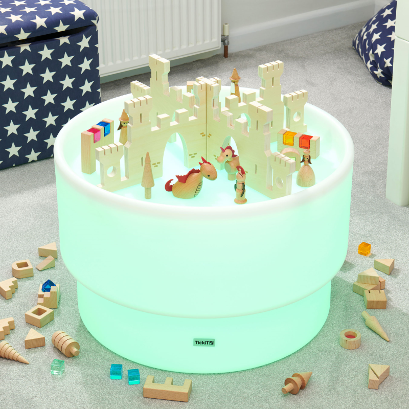 Toy castle set on a glowing green sensory  table with scattered wooden toys in a room setting.