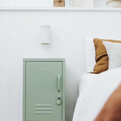 Green metal locker against a white wall with a bed and decorative items in the background