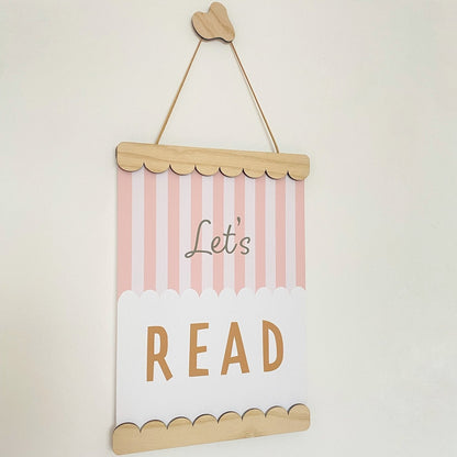 Decorative sign with 'Let's Read' text on a striped background, hanging against a light beige wall.